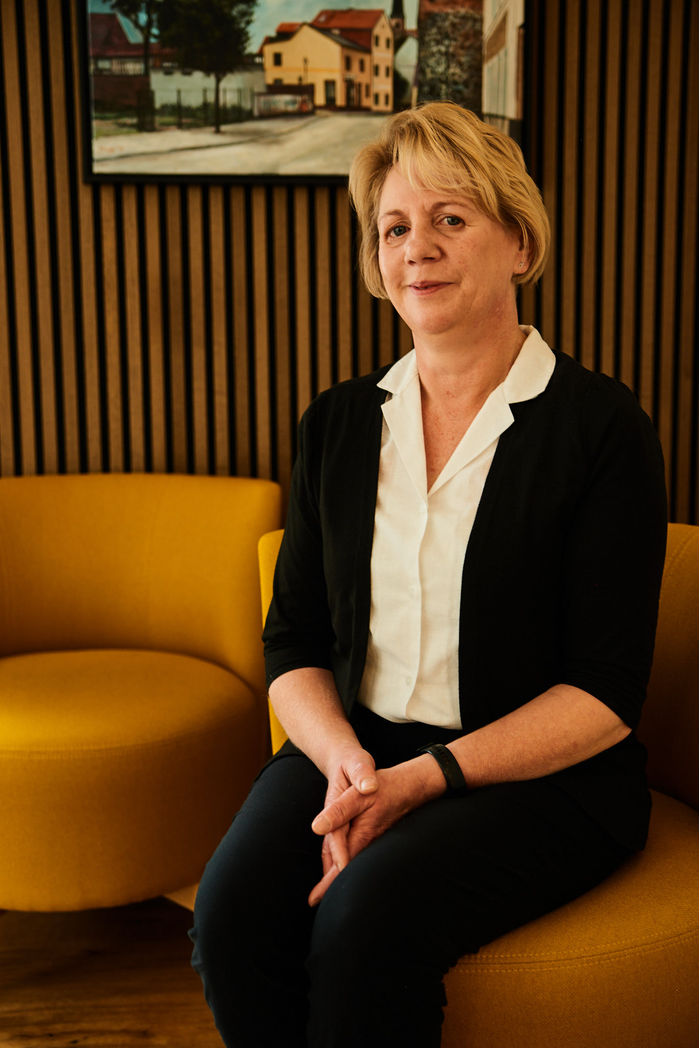 Eine Frau mit blondem Haar sitzt lächelnd auf einem gelben Sessel vor Holzpaneelen