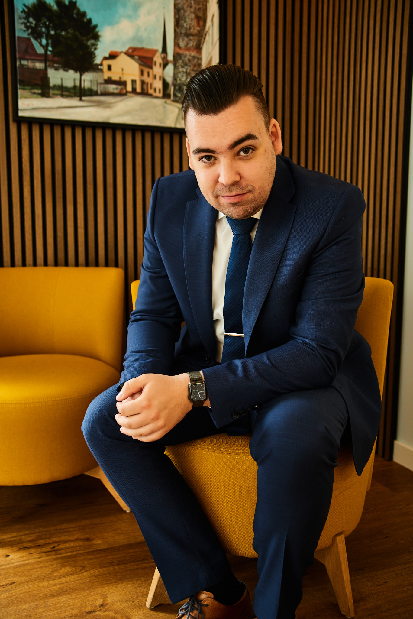 Ein junger Mann im blauen Anzug sitzt selbstbewusst auf einem gelben Sessel