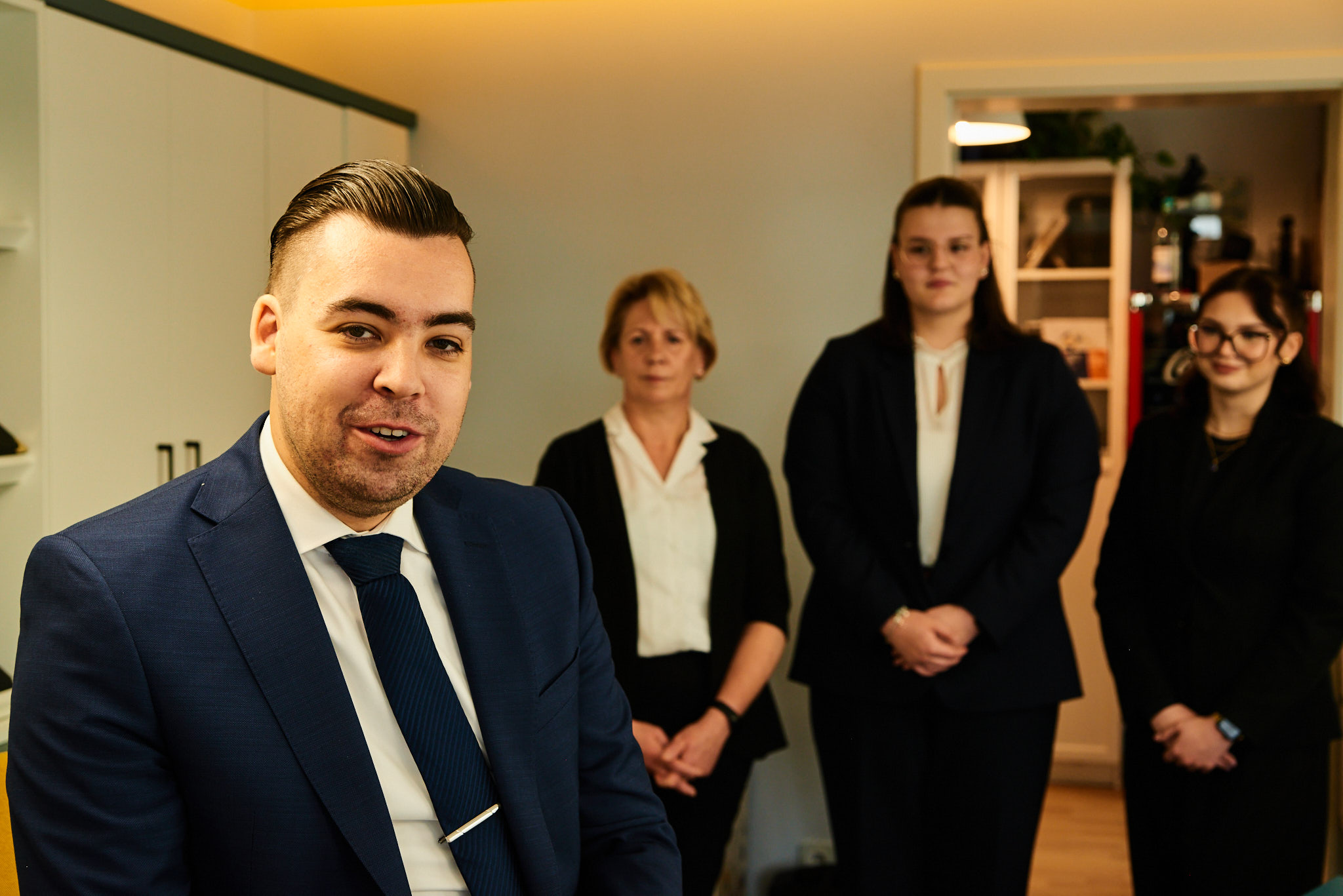 Ein junger Mann im blauen Anzug lächelt, drei Frauen stehen im Hintergrund im Büro