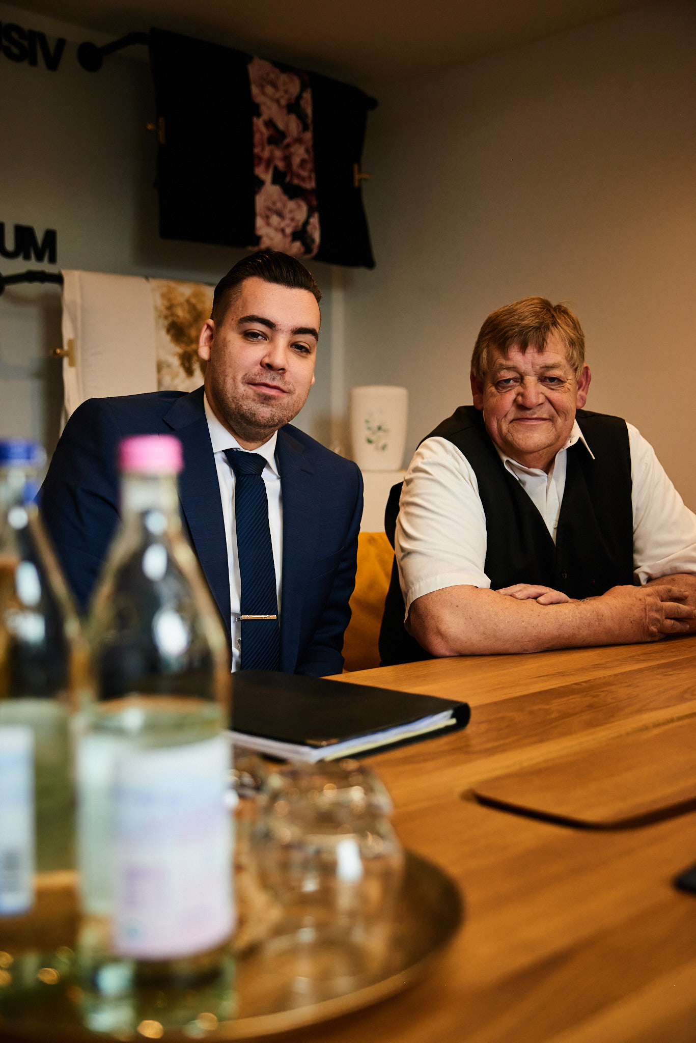 Zwei Männer sitzen freundlich lächelnd an einem Holztisch mit Wasserflaschen im Vordergrund