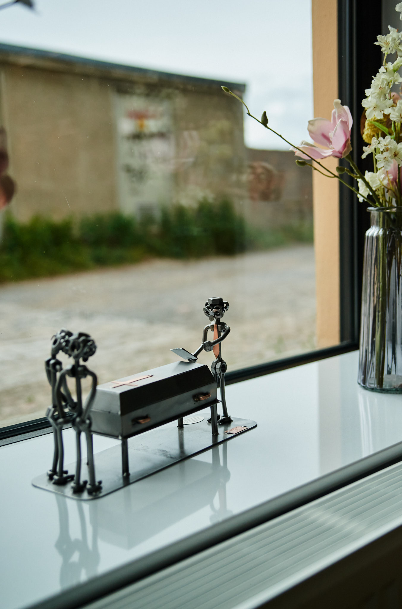 Eine silberne Metallfigurengruppe steht auf einem Fensterbrett vor einer Blumenvase