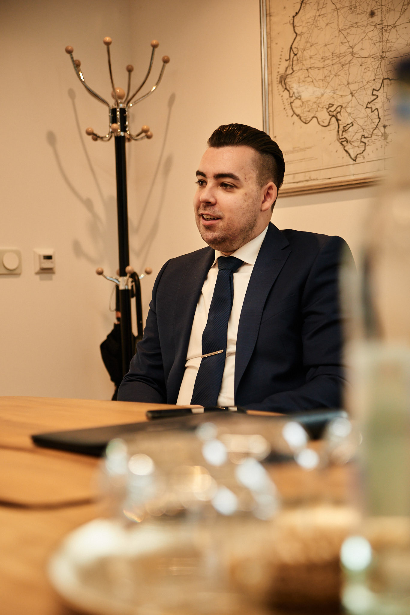Junger Mann im dunkelblauen Anzug sitzt lächelnd an einem Holztisch im Büro