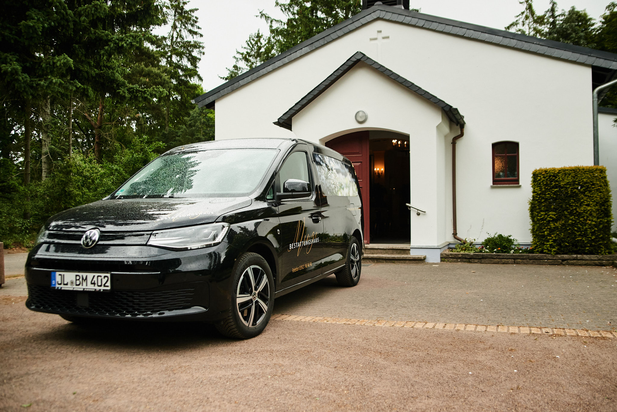 Schwarzer VW Van steht vor weißer Kapelle, Licht scheint aus der offenen Tür